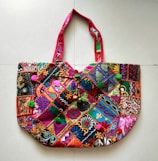 A richly embroidered handbag featuring a vibrant patchwork design. The fabric is adorned with intricate patterns and colors, including floral and geometric motifs. The handles are crafted with complementary red fabric, adding to the overall colorful theme. The bag is embellished with small pom-poms and beads, enhancing its decorative appeal.