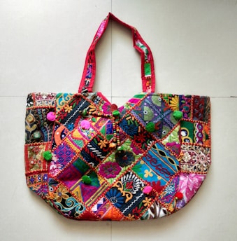 A richly embroidered handbag featuring a vibrant patchwork design. The fabric is adorned with intricate patterns and colors, including floral and geometric motifs. The handles are crafted with complementary red fabric, adding to the overall colorful theme. The bag is embellished with small pom-poms and beads, enhancing its decorative appeal.