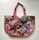 A richly embroidered handbag featuring a vibrant patchwork design. The fabric is adorned with intricate patterns and colors, including floral and geometric motifs. The handles are crafted with complementary red fabric, adding to the overall colorful theme. The bag is embellished with small pom-poms and beads, enhancing its decorative appeal.