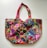 A richly embroidered handbag featuring a vibrant patchwork design. The fabric is adorned with intricate patterns and colors, including floral and geometric motifs. The handles are crafted with complementary red fabric, adding to the overall colorful theme. The bag is embellished with small pom-poms and beads, enhancing its decorative appeal.