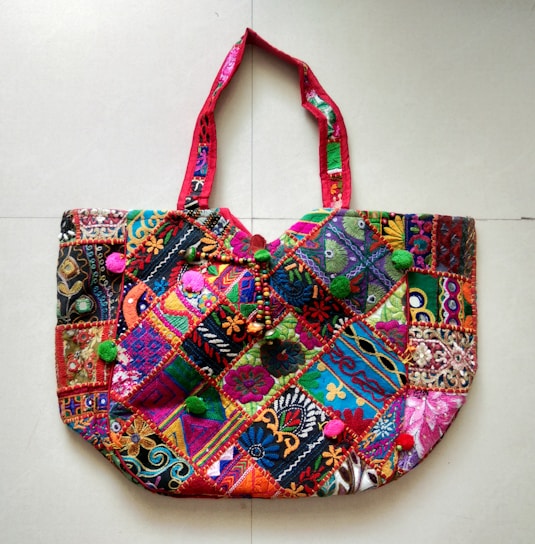 A richly embroidered handbag featuring a vibrant patchwork design. The fabric is adorned with intricate patterns and colors, including floral and geometric motifs. The handles are crafted with complementary red fabric, adding to the overall colorful theme. The bag is embellished with small pom-poms and beads, enhancing its decorative appeal.