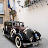A vintage black and brown car with white-wall tires is parked on a street next to a building with a Spanish flag. The car has classic chrome detailing and decorative flowers attached to the side mirror, suggesting it might be used for a special occasion. The background includes a street lamp and balconies with plants.