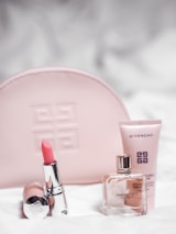 A collection of Givenchy beauty products is arranged neatly. A pink embossed makeup bag is in the background. In the foreground, a pink lipstick in a shiny silver tube is placed next to a clear glass perfume bottle labeled 'Irresistible Givenchy' and a pink tube of skincare product.