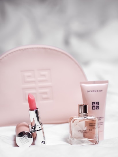 A collection of Givenchy beauty products is arranged neatly. A pink embossed makeup bag is in the background. In the foreground, a pink lipstick in a shiny silver tube is placed next to a clear glass perfume bottle labeled 'Irresistible Givenchy' and a pink tube of skincare product.