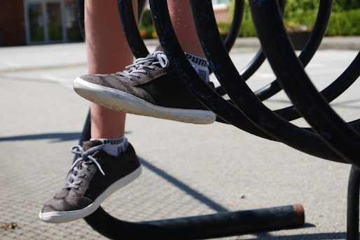 A pair of legs wearing sneakers positioned between the bars of a metallic spiral structure. The surfaces of the sneakers appear worn, suggesting active use, and there are shadows cast on the ground. The background includes blurred greenery and a building.