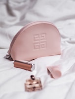 A pink cosmetic bag with a designer logo is surrounded by a lipstick, a small perfume bottle, and a tube of cream, all placed on a white quilted fabric.