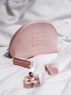 A pink cosmetic bag with a designer logo is surrounded by a lipstick, a small perfume bottle, and a tube of cream, all placed on a white quilted fabric.