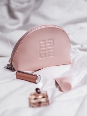 A pink cosmetic bag with a designer logo is surrounded by a lipstick, a small perfume bottle, and a tube of cream, all placed on a white quilted fabric.