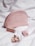 A pink cosmetic bag with a designer logo is surrounded by a lipstick, a small perfume bottle, and a tube of cream, all placed on a white quilted fabric.