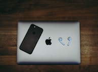 A black smartphone and a pair of wireless earbuds are placed on a closed silver laptop, all resting on a wooden surface. The phone and laptop display a prominent logo.