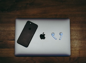 A black smartphone and a pair of wireless earbuds are placed on a closed silver laptop, all resting on a wooden surface. The phone and laptop display a prominent logo.