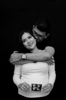 A couple is posing in an affectionate embrace. The man stands behind the woman, wrapping his arms around her shoulders. The woman is smiling, holding an ultrasound image in her hands, signifying pregnancy. The background is completely black, drawing focus to the couple.