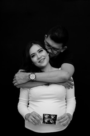 A couple is posing in an affectionate embrace. The man stands behind the woman, wrapping his arms around her shoulders. The woman is smiling, holding an ultrasound image in her hands, signifying pregnancy. The background is completely black, drawing focus to the couple.