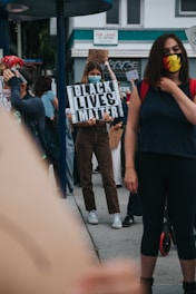 Several individuals are gathered outside with some holding signs advocating for the Black Lives Matter movement. One sign prominently reads 'Black Lives Matter'. The participants are wearing face masks, suggesting a concern for health safety. The setting appears to be an urban environment, with buildings and a 'For Lease' sign visible in the background.