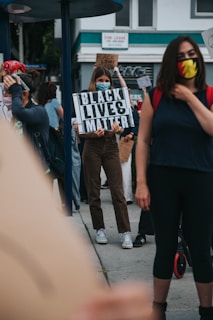 Several individuals are gathered outside with some holding signs advocating for the Black Lives Matter movement. One sign prominently reads 'Black Lives Matter'. The participants are wearing face masks, suggesting a concern for health safety. The setting appears to be an urban environment, with buildings and a 'For Lease' sign visible in the background.