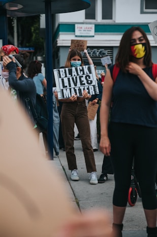 Several individuals are gathered outside with some holding signs advocating for the Black Lives Matter movement. One sign prominently reads 'Black Lives Matter'. The participants are wearing face masks, suggesting a concern for health safety. The setting appears to be an urban environment, with buildings and a 'For Lease' sign visible in the background.