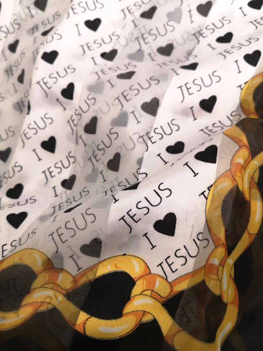 A fabric designed with repeating text 'I ❤️ JESUS' interspersed with black heart icons. A decorative pattern with yellow and orange chains is visible at the lower corner.