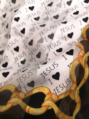 A fabric designed with repeating text 'I ❤️ JESUS' interspersed with black heart icons. A decorative pattern with yellow and orange chains is visible at the lower corner.