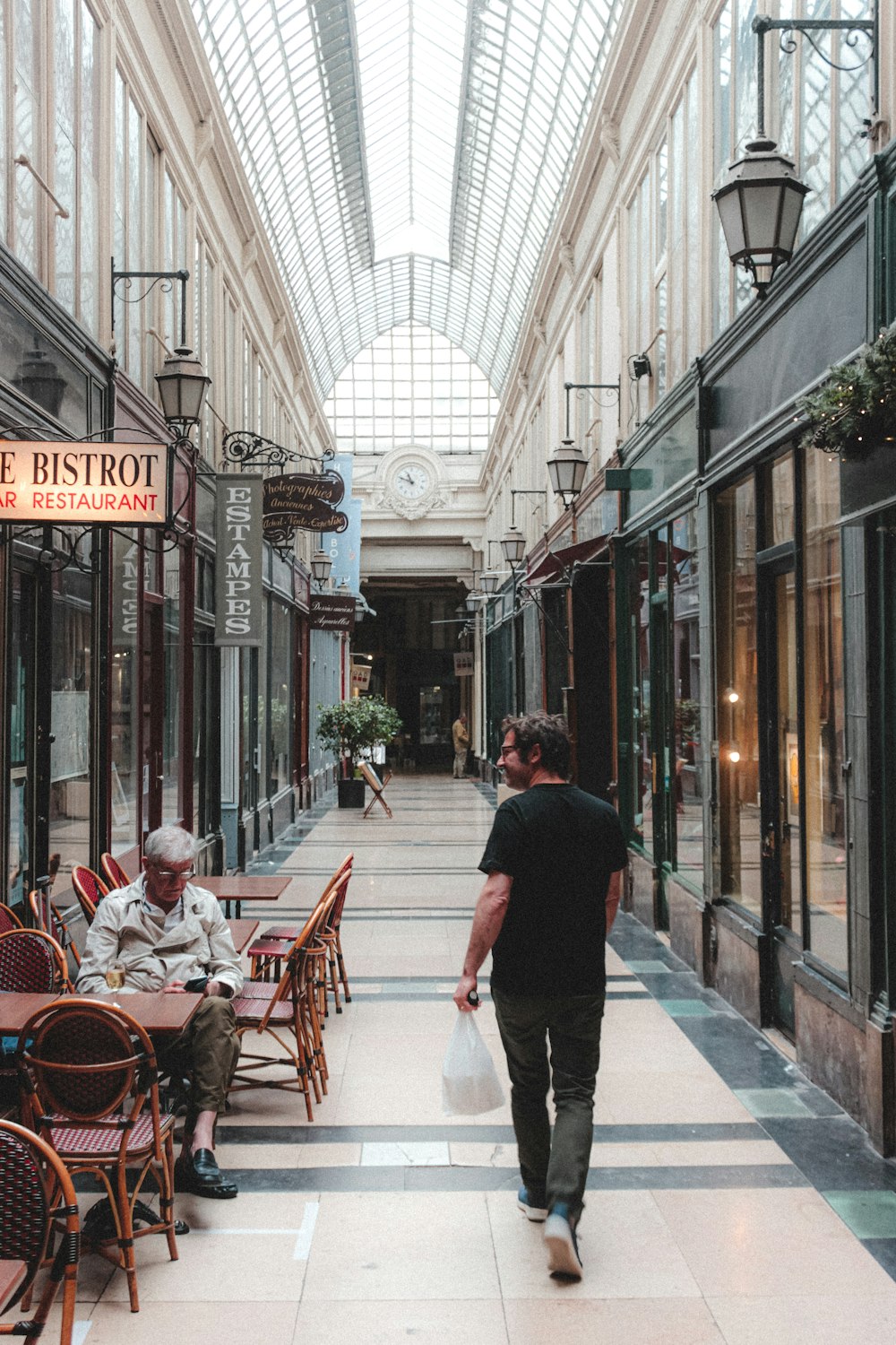 man in black jacket and white pants holding white plastic bag walking on sidewalk during daytime