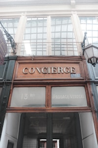 An old-fashioned entrance with a sign reading 'CONCIERGE' above a wooden-framed doorway. The upper part of the entrance displays the address '13 BIS' and 'Passage Verdeau'. The architecture is classic with decorative elements and a large glass paneling at the top. A vintage lantern is hanging on the right.