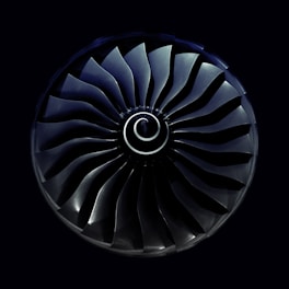 A symmetrical view of a jet engine turbine with curved blades radiating from a central spiral hub, set against a dark background.