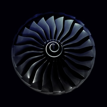 A symmetrical view of a jet engine turbine with curved blades radiating from a central spiral hub, set against a dark background.