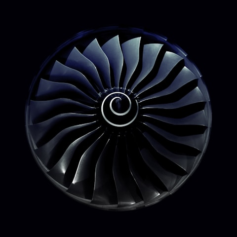 A symmetrical view of a jet engine turbine with curved blades radiating from a central spiral hub, set against a dark background.