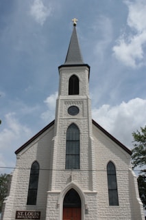 A historic church building with a tall, pointed steeple topped by a cross. The facade is made of textured white stone, featuring arched windows with intricate designs. The entrance is marked by a large arched door. A sign near the entrance reads 'St. Louis Besancon'. The sky is partly cloudy, providing a serene backdrop.