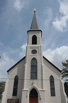 A historic church building with a tall, pointed steeple topped by a cross. The facade is made of textured white stone, featuring arched windows with intricate designs. The entrance is marked by a large arched door. A sign near the entrance reads 'St. Louis Besancon'. The sky is partly cloudy, providing a serene backdrop.