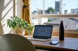 A wooden desk positioned by a window with a view of buildings and a church tower. On the desk are a green plant in a white pot, an open laptop displaying a document, a black water bottle, and a smartphone. Yellow curtains are partially drawn to the sides of the window, allowing natural light to fill the room.