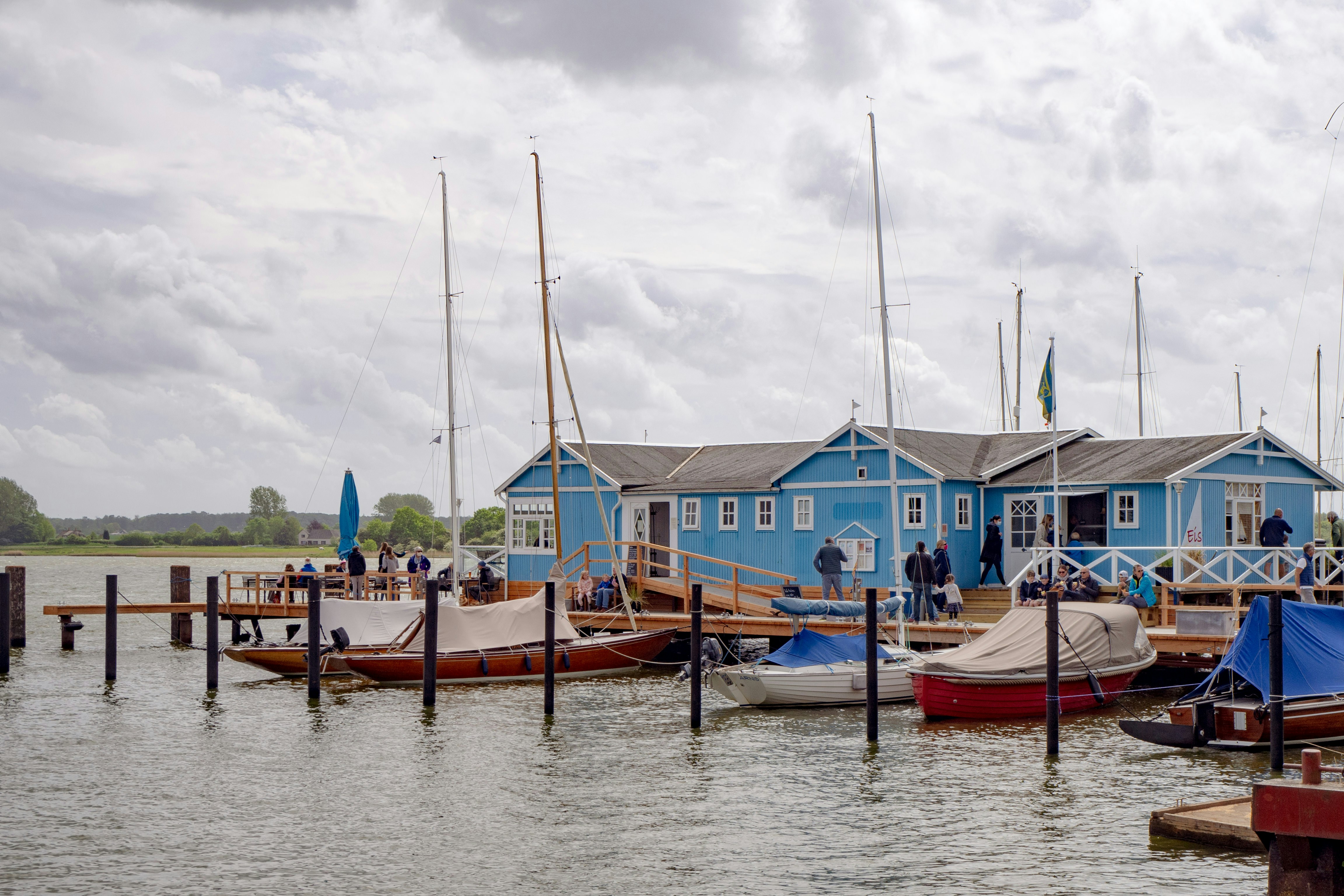 Menschen unterhalten sich auf einem Holzsteg in der Nähe eines blauen Bootshauses.