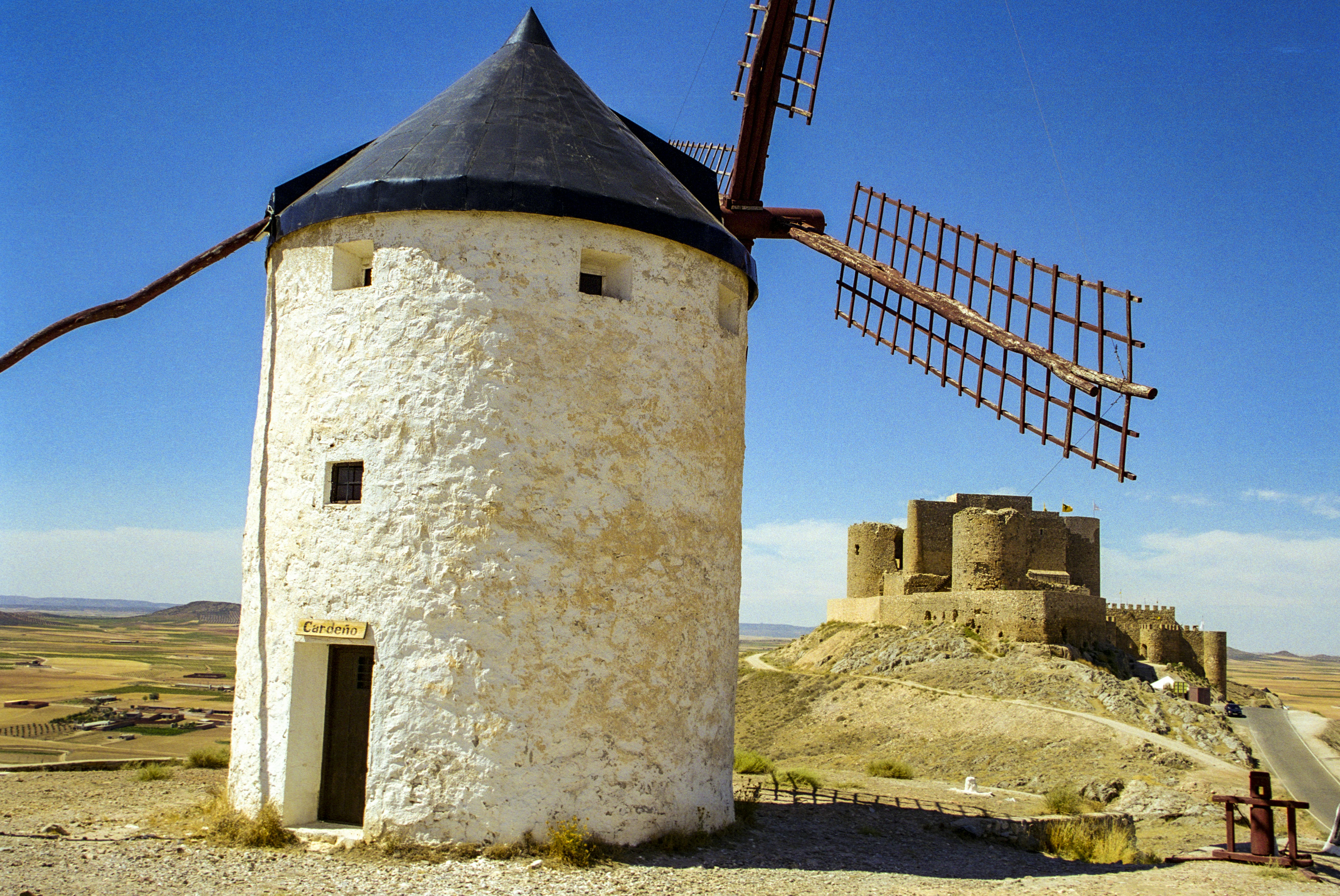 brown brick building near red metal tower, A row of 12 windmills line a hillside ridge to the south of Consuegra, Spain. Built in the 19th century to grind flour, they are the local favorite for watching the sunset. The Castillo de Consuegra has great views and a bird of prey exhibit. It makes for an interesting history lesson south of Madrid on the way to Cordoba. 