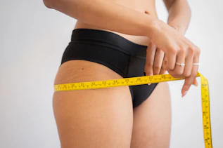 A person in black underwear measuring their hip with a yellow tape measure