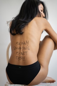 A person sits on a chair wearing black underwear, with long dark hair covering part of their face. The phrase 'Skinny shaming is not okay' is written on their back.