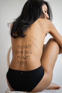 A person sits on a chair wearing black underwear, with long dark hair covering part of their face. The phrase 'Skinny shaming is not okay' is written on their back.