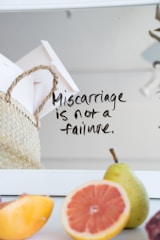 A handwritten note on a mirror reads 'Miscarriage is not a failure.' A woven basket containing papers and an assortment of fresh fruits, including a pear, grapefruit, and peach are visible. The background is softly blurred.