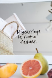 A handwritten note on a mirror reads 'Miscarriage is not a failure.' A woven basket containing papers and an assortment of fresh fruits, including a pear, grapefruit, and peach are visible. The background is softly blurred.