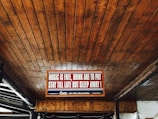 A wooden ceiling with an illuminated sign that reads 'Music is free, drink are to pay, stay till late but sleep away!' with a decorative border. The sign is centrally placed and accompanied by subdued lighting.