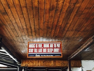 A wooden ceiling with an illuminated sign that reads 'Music is free, drink are to pay, stay till late but sleep away!' with a decorative border. The sign is centrally placed and accompanied by subdued lighting.