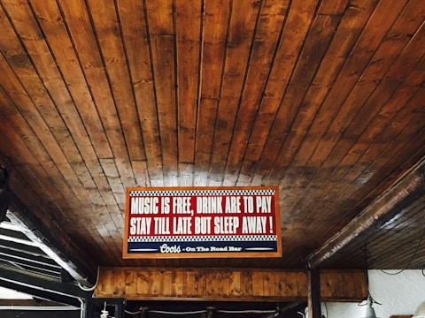 A wooden ceiling with an illuminated sign that reads 'Music is free, drink are to pay, stay till late but sleep away!' with a decorative border. The sign is centrally placed and accompanied by subdued lighting.