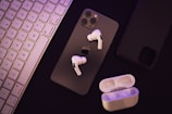 A set of wireless earphones placed over a smartphone with a visible Apple logo. Beside them, there is a charging case for the earphones. A keyboard can be seen to the left, and another smartphone appears on the right.