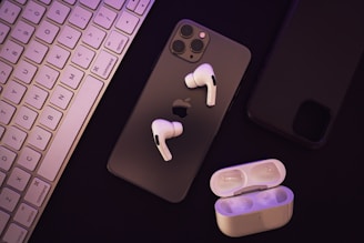 A set of wireless earphones placed over a smartphone with a visible Apple logo. Beside them, there is a charging case for the earphones. A keyboard can be seen to the left, and another smartphone appears on the right.
