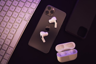 A set of wireless earphones placed over a smartphone with a visible Apple logo. Beside them, there is a charging case for the earphones. A keyboard can be seen to the left, and another smartphone appears on the right.