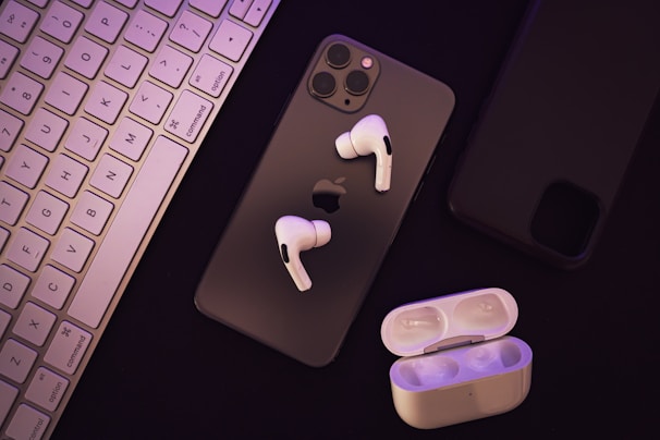 A set of wireless earphones placed over a smartphone with a visible Apple logo. Beside them, there is a charging case for the earphones. A keyboard can be seen to the left, and another smartphone appears on the right.