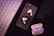 A set of wireless earphones placed over a smartphone with a visible Apple logo. Beside them, there is a charging case for the earphones. A keyboard can be seen to the left, and another smartphone appears on the right.