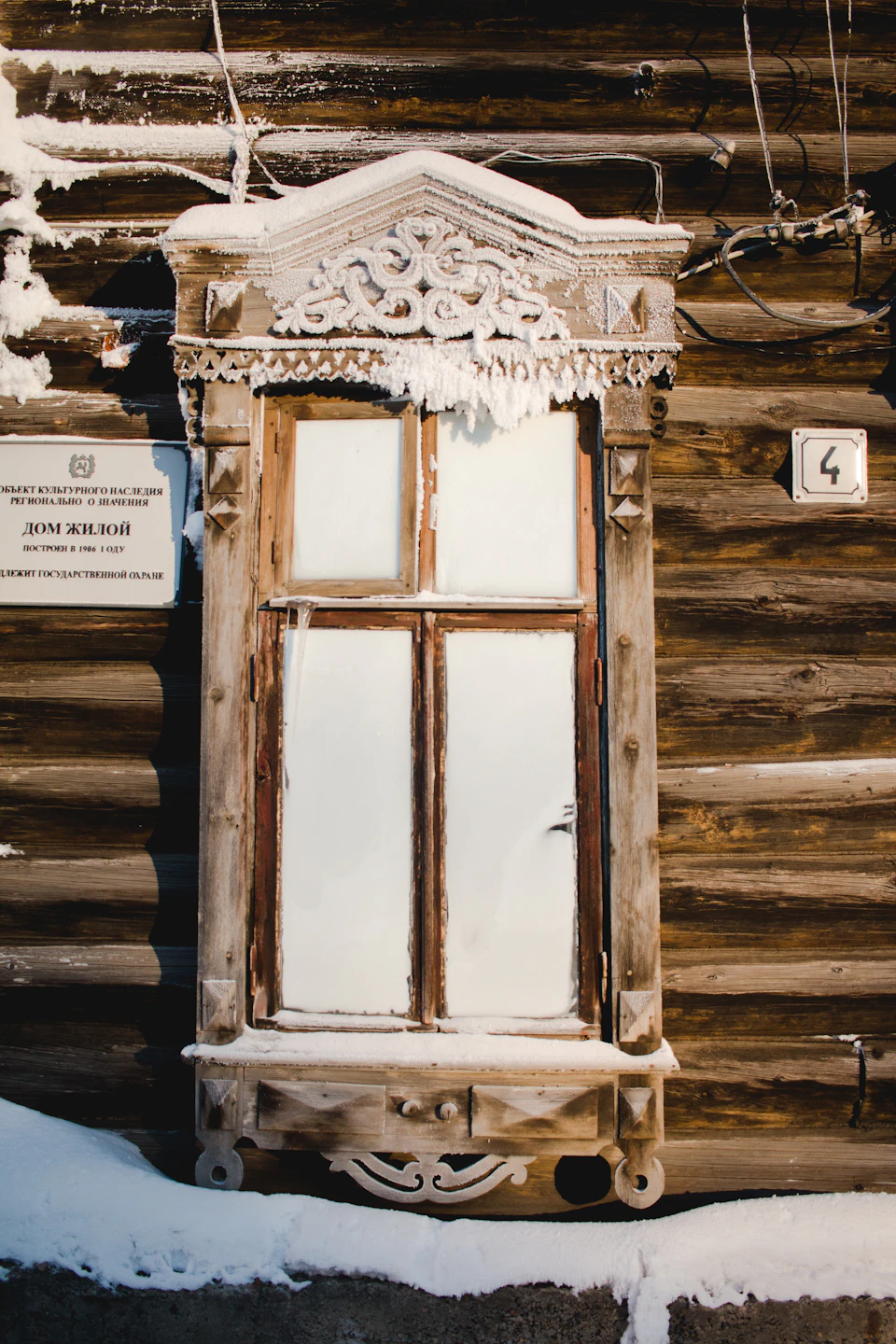 Wooden building with ornate window partially covered in snow and a sign attached nearby