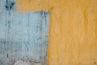 A textured wall with two distinct sections, one painted in a faded blue and the other in a mustard yellow. The dividing line between the colors is irregular and there are visible drips and smudges creating an abstract appearance.