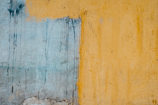 A textured wall with two distinct sections, one painted in a faded blue and the other in a mustard yellow. The dividing line between the colors is irregular and there are visible drips and smudges creating an abstract appearance.