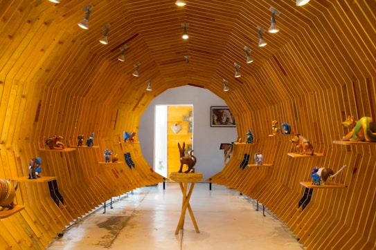 A gallery space designed with a tunnel-like wooden structure, showcasing a variety of colorful, artistic figurines displayed on small shelves along the walls. The ceiling is adorned with spotlights that illuminate the interior. At the end of the tunnel, natural light enters through an open door, adding warmth to the setting.