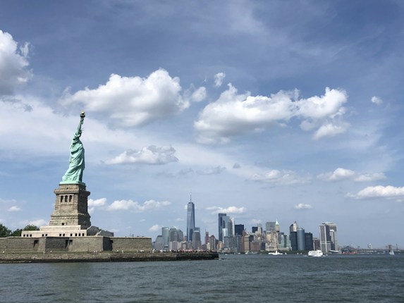 A view of the Statue of Liberty on Liberty Island with the New York City skyline in the background under a blue sky with scattered clouds.